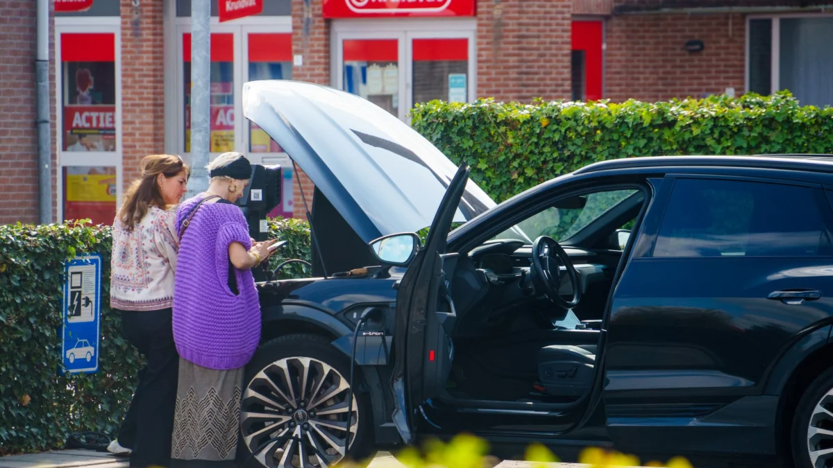 Twee vrouwen staan naast een auto met een open motorkap voor een winkel, mogelijk om problemen te controleren.