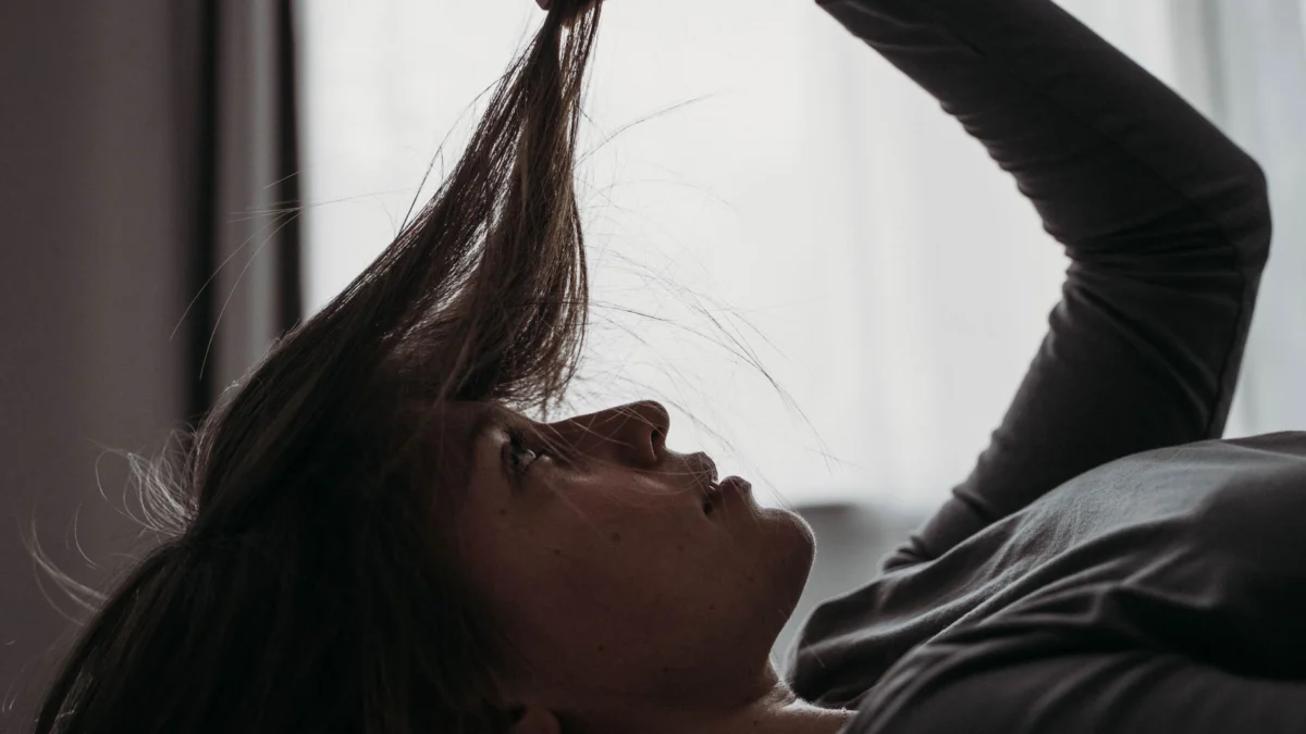 Zijaanzicht van een vrouw die in bed ligt en haar haar bedachtzaam vasthoudt, binnenshuis.