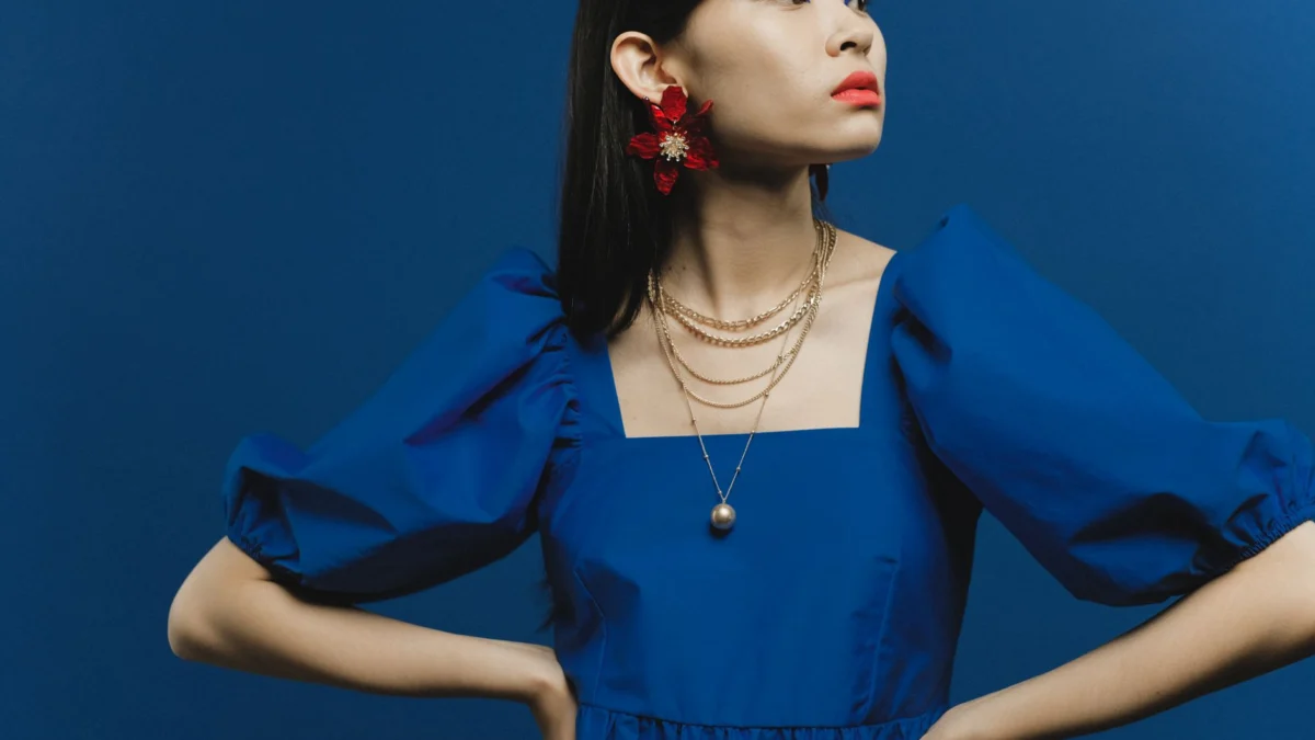 Aziatische vrouw die poseert in een levendige blauwe jurk met gedurfde make-up en rode bloemenoorbellen.