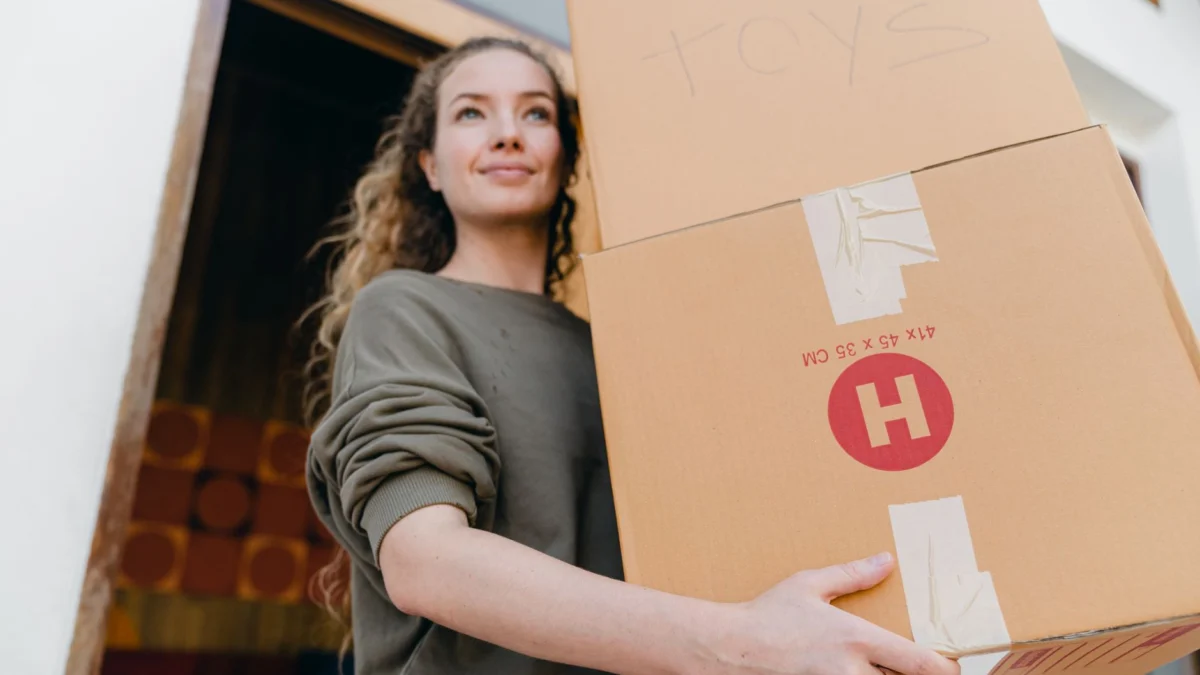 Jonge vrouw draagt zelfverzekerd verhuisdozen buiten, wat verhuis of verandering van levensstijl aangeeft.