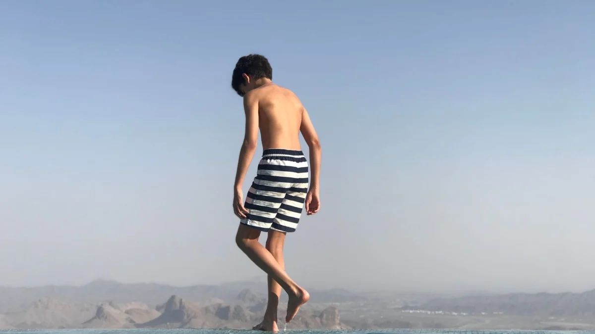 Een jongen in gestreepte zwemshorts geniet van een zonnige dag bij een infinity pool met uitzicht op de bergen.