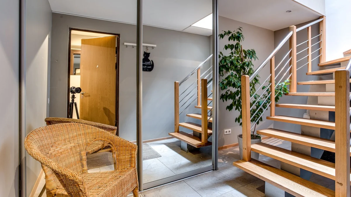 Moderne woninginrichting met een glazen kledingkast, houten trap, potplant en rieten stoel.
