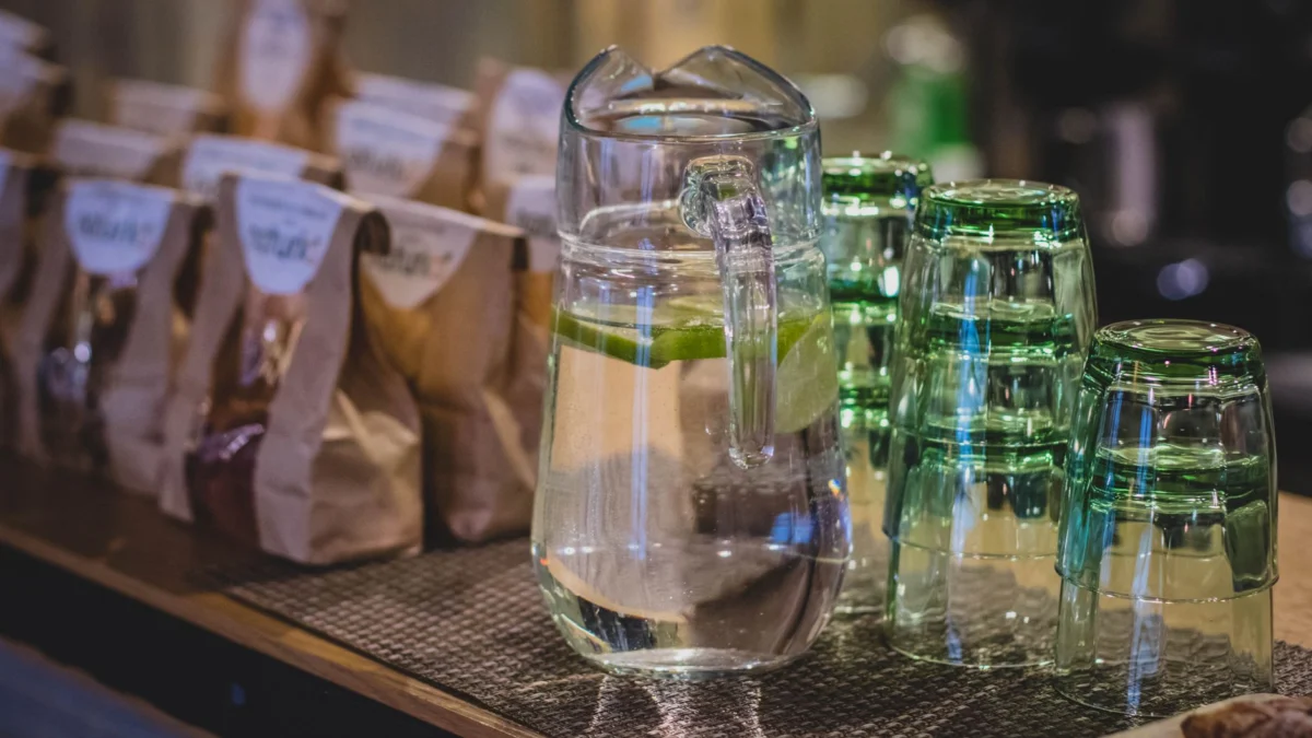 Een glazen kan gevuld met limoenwater staat op de toonbank van een koffiebar, versierd met opgestapelde glazen en koffietassen.